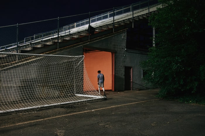 Teenage male in sportswear stands in an orange-lit tunnel entrance to a stadium; a soccer net rests in the foreground.
