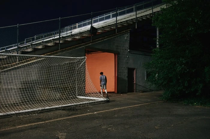 Teenage male in sportswear stands in an orange-lit tunnel entrance to a stadium; a soccer net rests in the foreground.