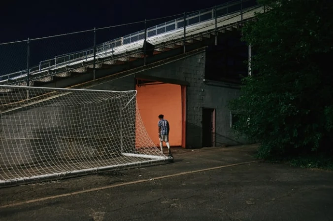 Teenage male in sportswear stands in an orange-lit tunnel entrance to a stadium; a soccer net rests in the foreground.