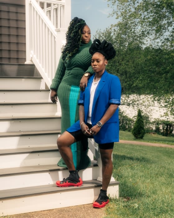 Two African American women pose on sunlit porch steps. One wears green knit dress, the other, a blue blazer with red-soled sneakers.
