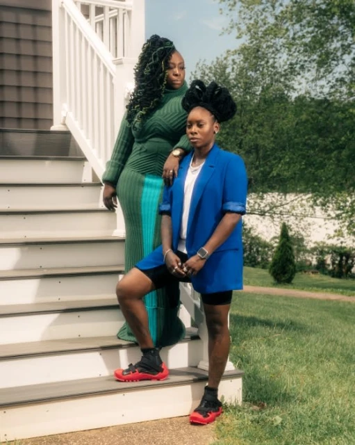 Two African American women pose on sunlit porch steps. One wears green knit dress, the other, a blue blazer with red-soled sneakers.