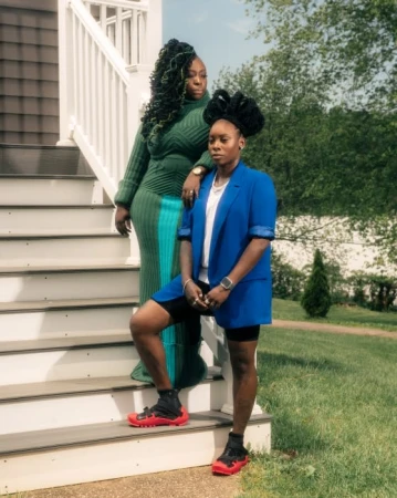 Two African American women pose on sunlit porch steps. One wears green knit dress, the other, a blue blazer with red-soled sneakers.