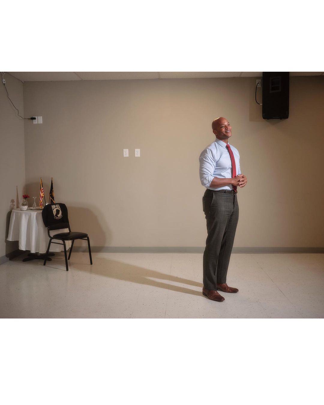Adult male stands in an indoor setting, wearing a light blue collared shirt, a red tie, and dark pants, near a small table.