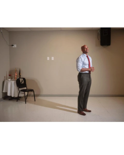Adult male stands in an indoor setting, wearing a light blue collared shirt, a red tie, and dark pants, near a small table.