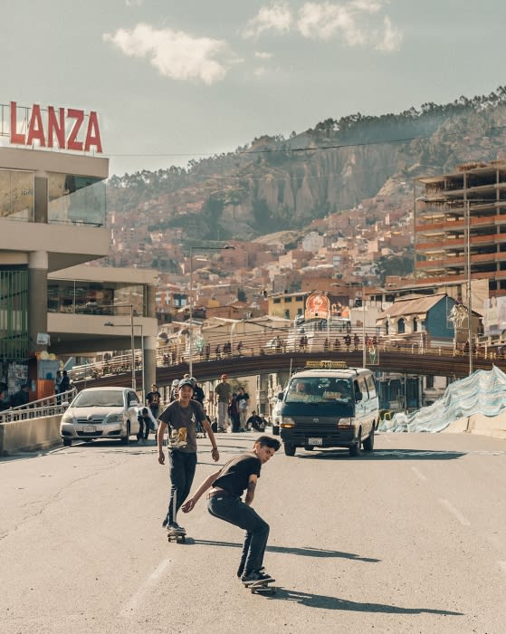 Two teenage boys ride skateboards down an urban street with a hillside city in the background under a bright blue sky.