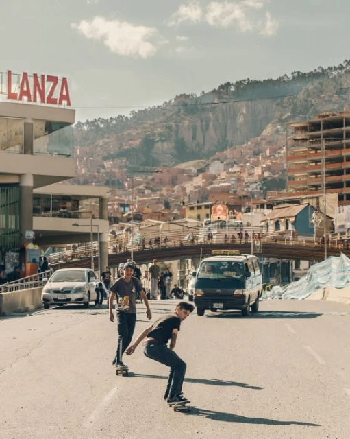 Two teenage boys ride skateboards down an urban street with a hillside city in the background under a bright blue sky.