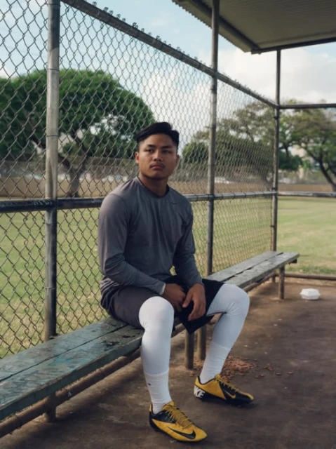 An adult male baseball player is sitting on a bench under a metal awning at a baseball field, wearing sportswear and cleats.