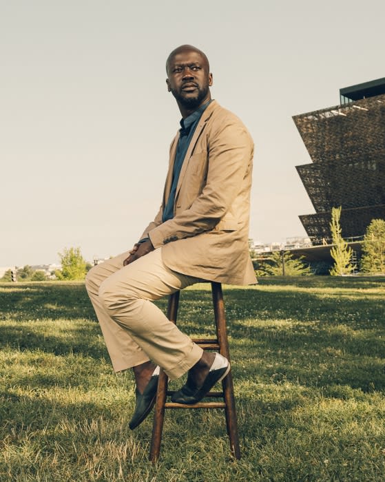 Man in beige suit sitting on stool outdoors with modern architecture. Low-angle shot of a person on a sunny day in a grassy area.