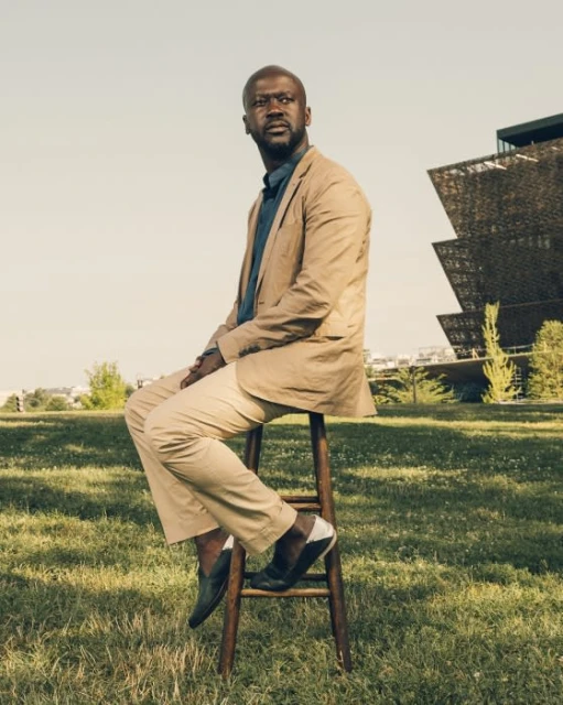 Man in beige suit sitting on stool outdoors with modern architecture. Low-angle shot of a person on a sunny day in a grassy area.