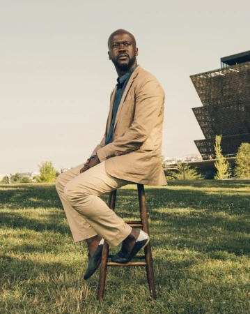 Man in beige suit sitting on stool outdoors with modern architecture. Low-angle shot of a person on a sunny day in a grassy area.