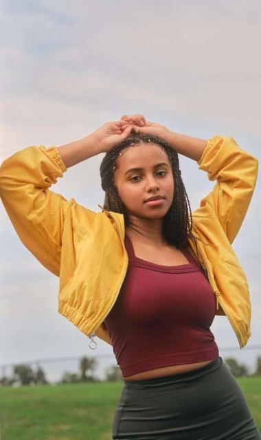 Portrait of a young woman with dark hair and yellow jacket in a field under a cloudy sky. She poses with arms above her head.