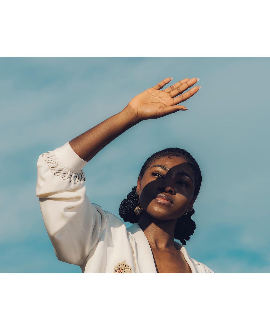 Woman with dark hair and fair skin stands outside on a sunny day, shielding her face with her hand, wearing a white jacket against a blue sky.