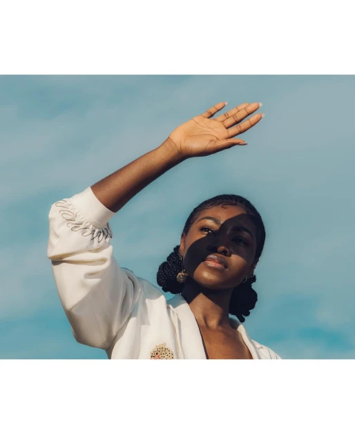 Woman with dark hair and fair skin stands outside on a sunny day, shielding her face with her hand, wearing a white jacket against a blue sky.