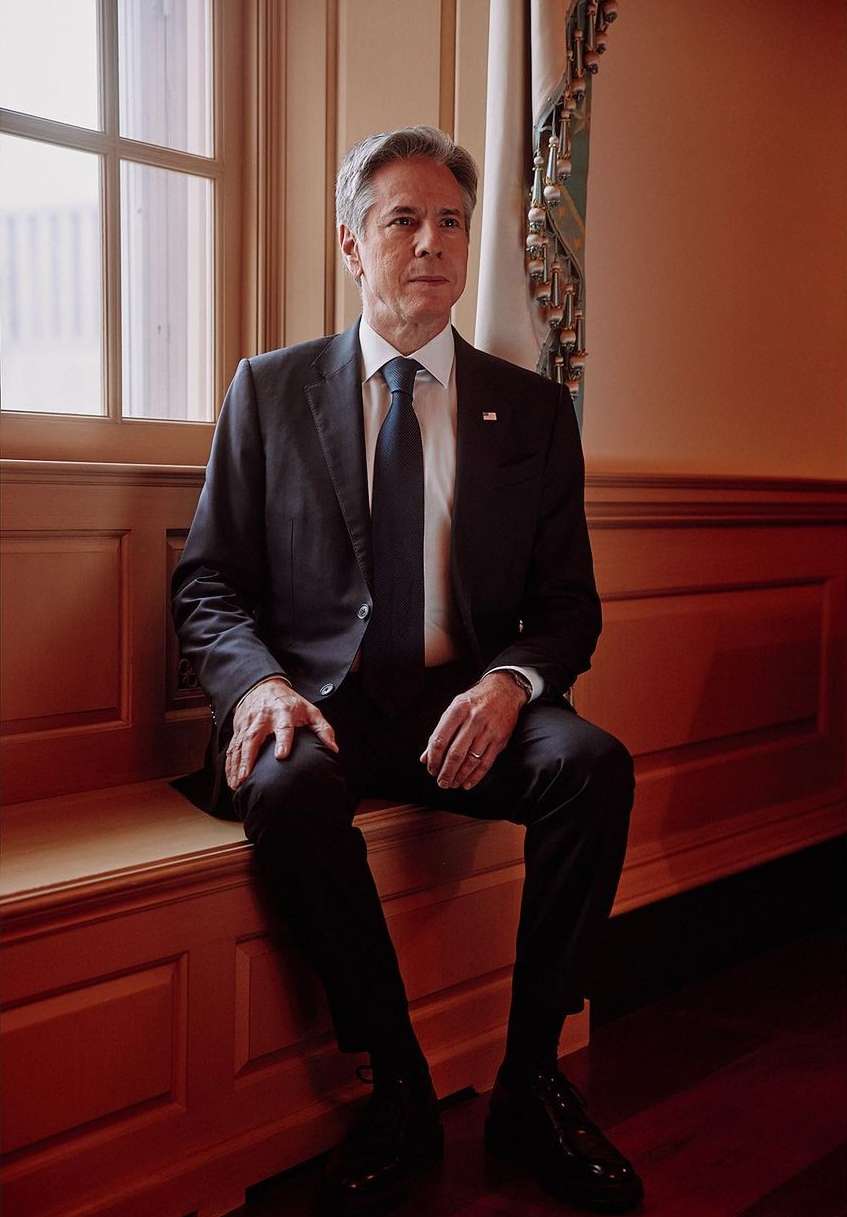 Portrait of a seated man in a suit near a window with natural light, wearing dark suit and tie with an American flag lapel pin.