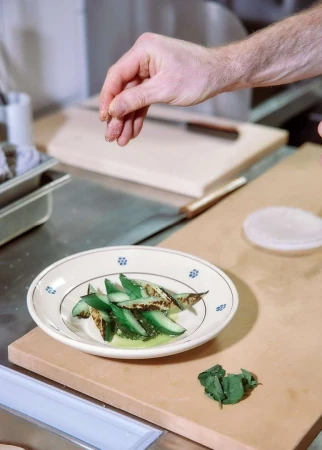 Chef's hand sprinkles seasoning on a plate of sliced grilled cucumber, plated with sprigs of herbs in a professional kitchen setting.
