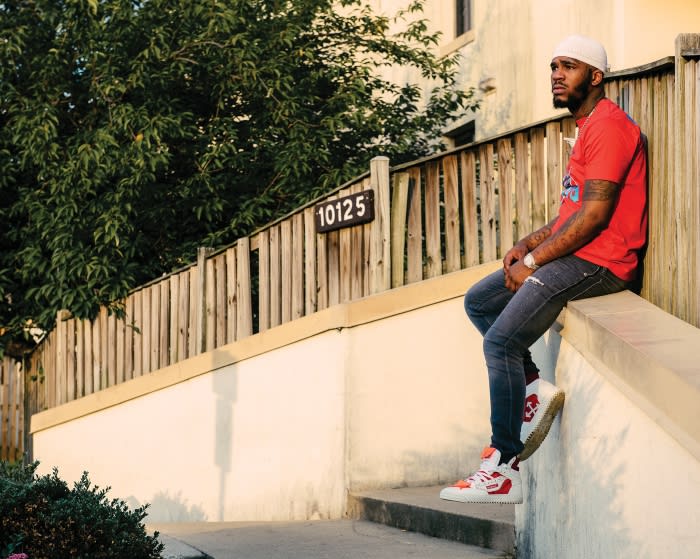 Man sitting on concrete ledge wearing a red t-shirt, jeans, white sneakers and a white hat in a residential neighborhood.