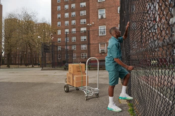A man in a teal outfit climbs a chain-link fence with boxes on a hand truck nearby, set against a backdrop of brick apartment buildings.
