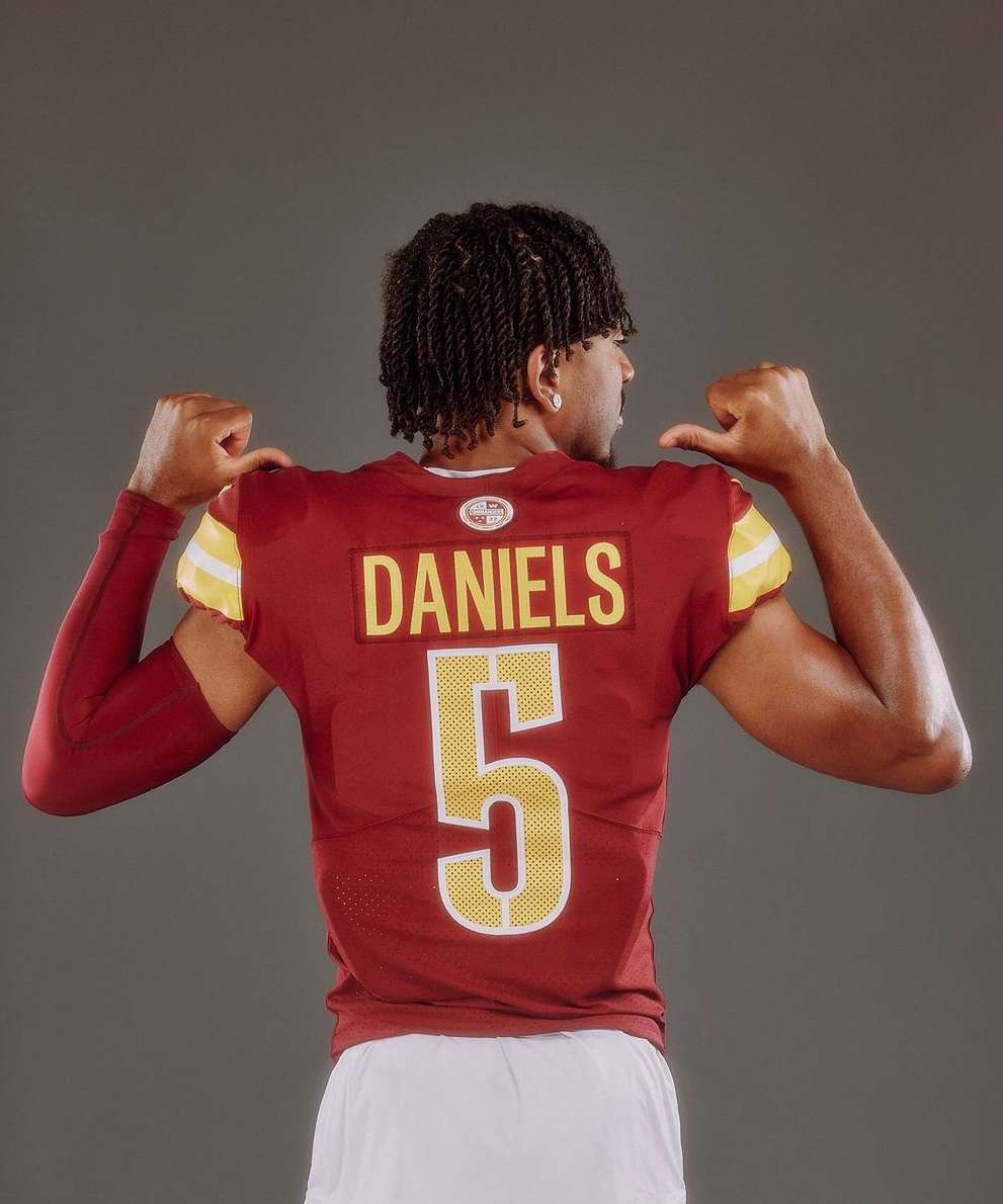 A male athlete with dark, braided hair poses in a maroon and gold football jersey with the number 5 on the back in a studio shot.
