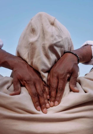 Close-up shot of a dark-skinned adult with their hooded head turned away from the camera, hands clasped behind their head against a sky-blue backdrop.