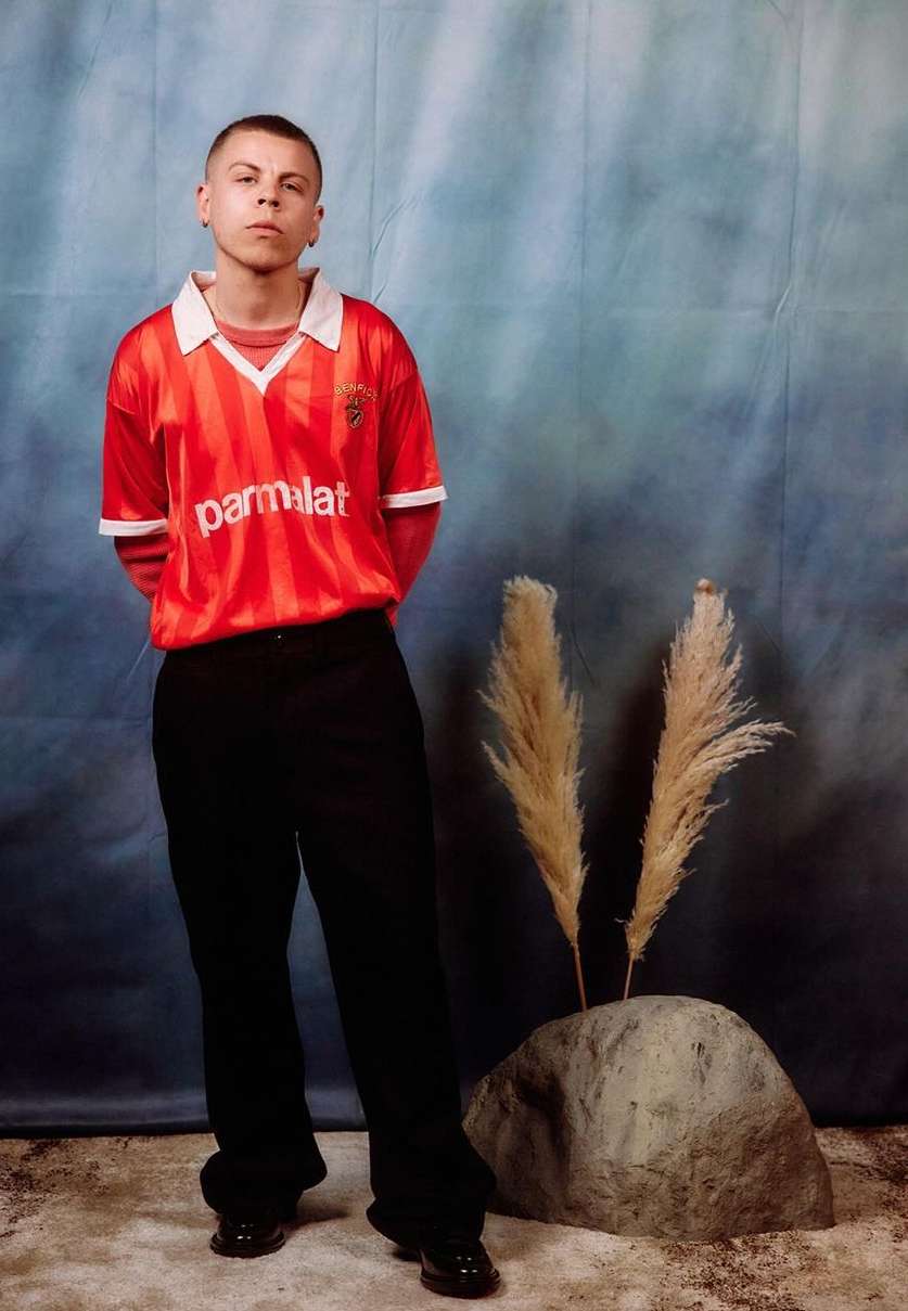Full-length portrait of a young adult male in a red soccer jersey and black pants, posing in front of a mottled blue backdrop with pampas grass.