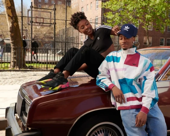A young woman sits on the hood of a car while a man leans against it, photographed by Jared Soares in an urban street setting.