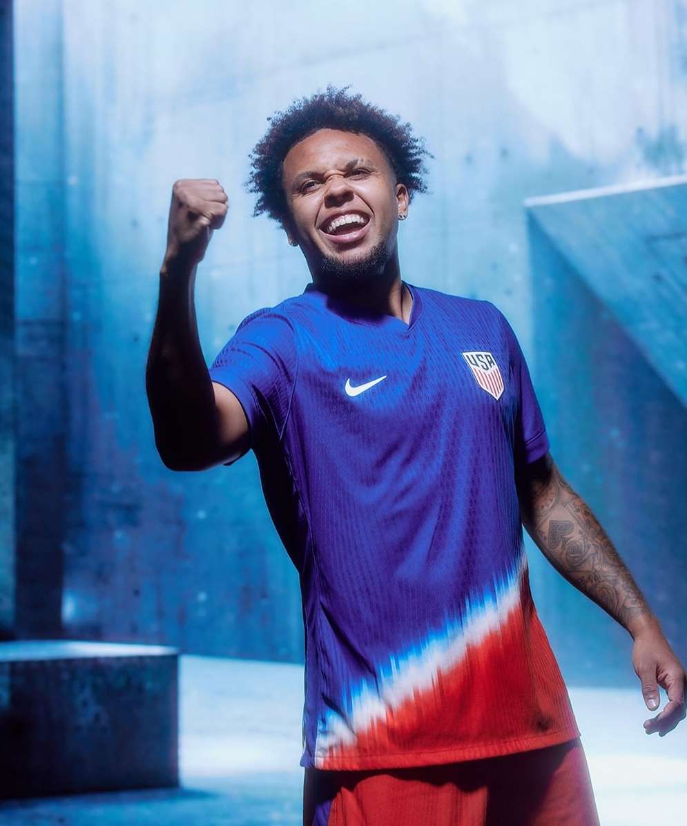 A man in a soccer jersey with a USA logo on it is shown from the waist up, with an excited expression and fist in the air.