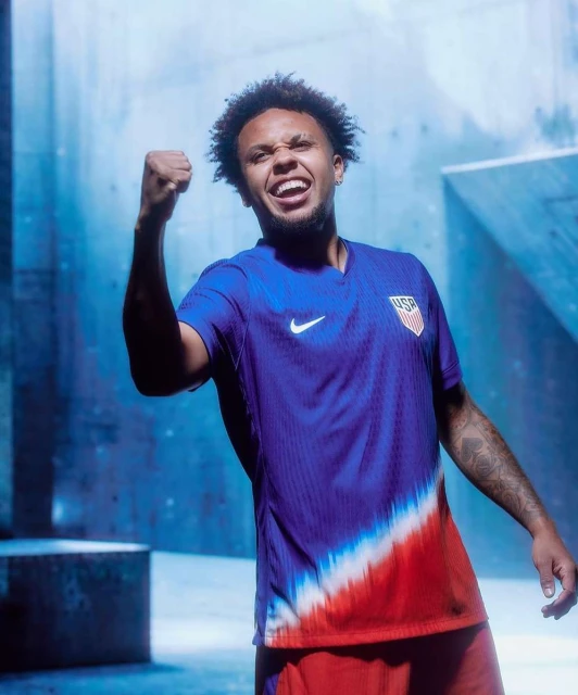 A man in a soccer jersey with a USA logo on it is shown from the waist up, with an excited expression and fist in the air.