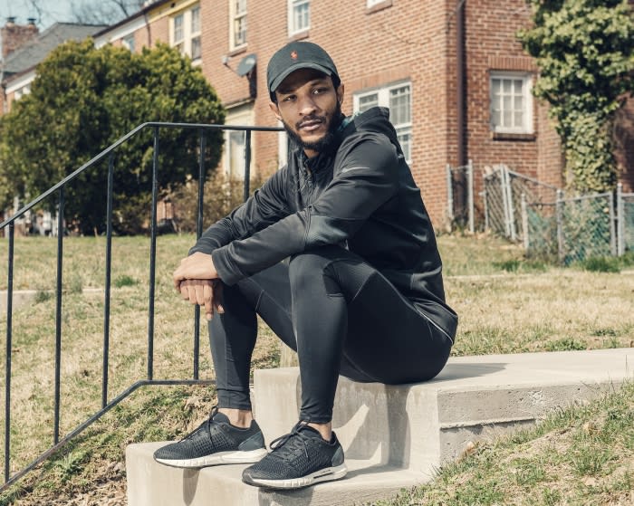 Adult male in athletic wear sits on concrete steps outdoors, wearing a cap and sneakers, with a brick building and green grass in background.