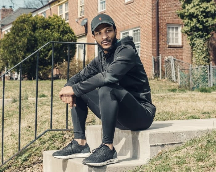Adult male in athletic wear sits on concrete steps outdoors, wearing a cap and sneakers, with a brick building and green grass in background.