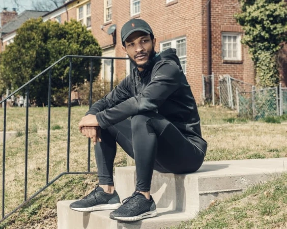Adult male in athletic wear sits on concrete steps outdoors, wearing a cap and sneakers, with a brick building and green grass in background.