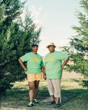 Two senior African-American women in matching green t-shirts hold hands in a sunlit rural landscape with trees and cloudy sky.
