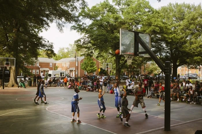 Outdoor basketball game on a gray court surrounded by a crowd, with trees, apartment buildings, and parked cars in the background.