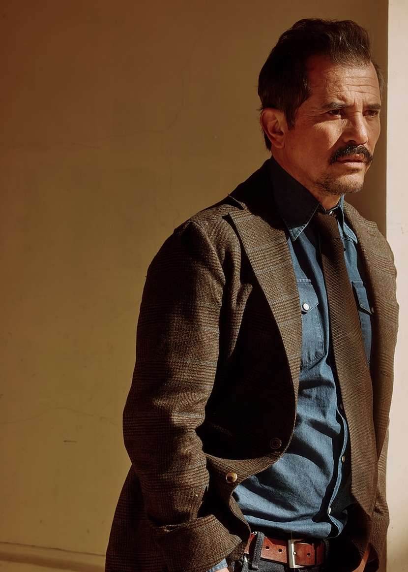 Portrait of a middle-aged man with a mustache wearing a brown jacket, tie, and blue denim shirt standing in front of a wall.