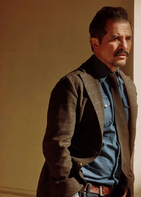 Portrait of a middle-aged man with a mustache wearing a brown jacket, tie, and blue denim shirt standing in front of a wall.