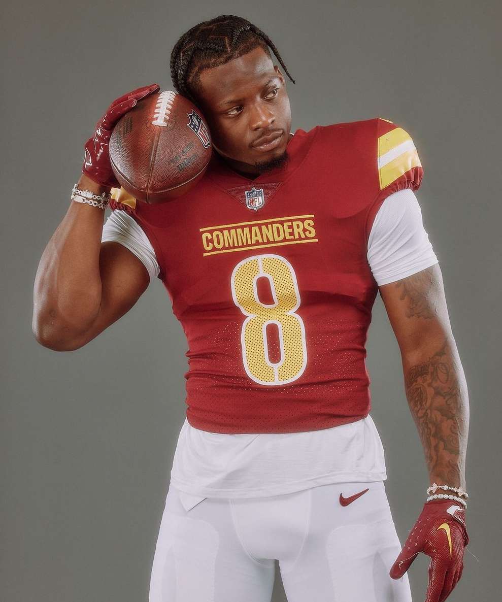 Football player in uniform poses with football in a studio, showcasing team colors of burgundy, gold, and white against a grey background.