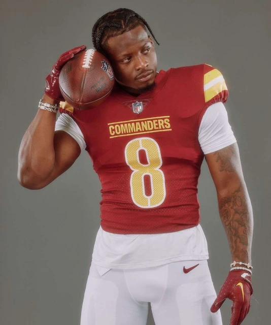 Football player in uniform poses with football in a studio, showcasing team colors of burgundy, gold, and white against a grey background.
