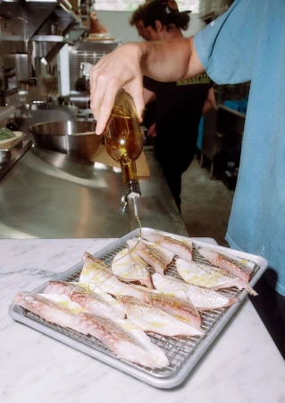 Fish fillets on a metal tray are drizzled with olive oil by a person in a kitchen setting, preparing for cooking or serving.