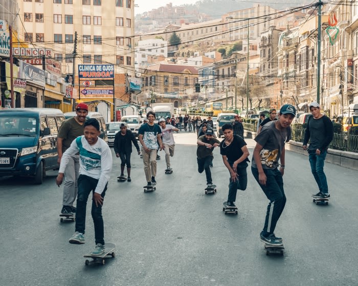 Group of teenagers riding skateboards on a city street lined with buildings and cars, capturing urban youth culture and active lifestyles.
