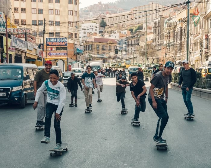 Group of teenagers riding skateboards on a city street lined with buildings and cars, capturing urban youth culture and active lifestyles.