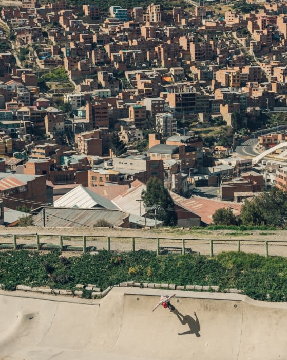 Skateboarder in a skatepark performs a trick, with an expansive view of a densely packed urban cityscape in the background during daylight.