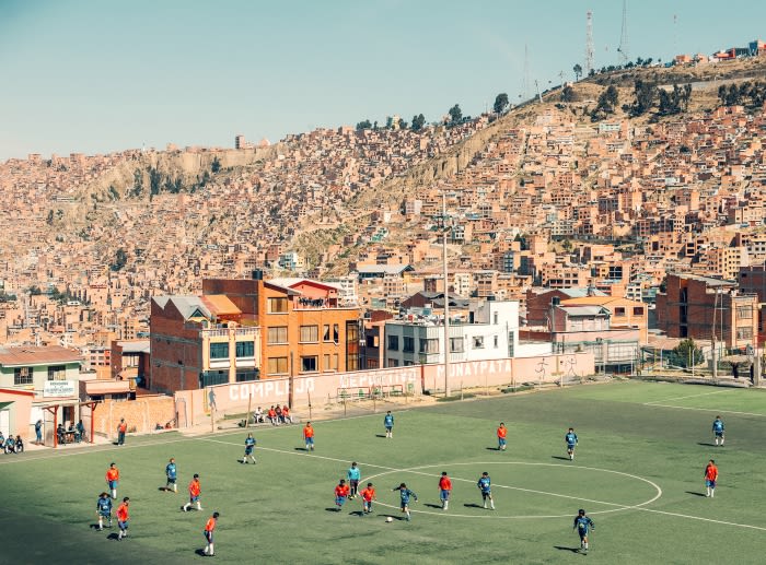Soccer players compete on a green field surrounded by buildings in La Paz, Bolivia. A densely packed hillside of houses rises behind.