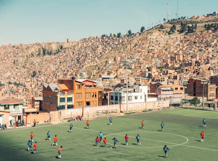 Soccer players compete on a green field surrounded by buildings in La Paz, Bolivia. A densely packed hillside of houses rises behind.