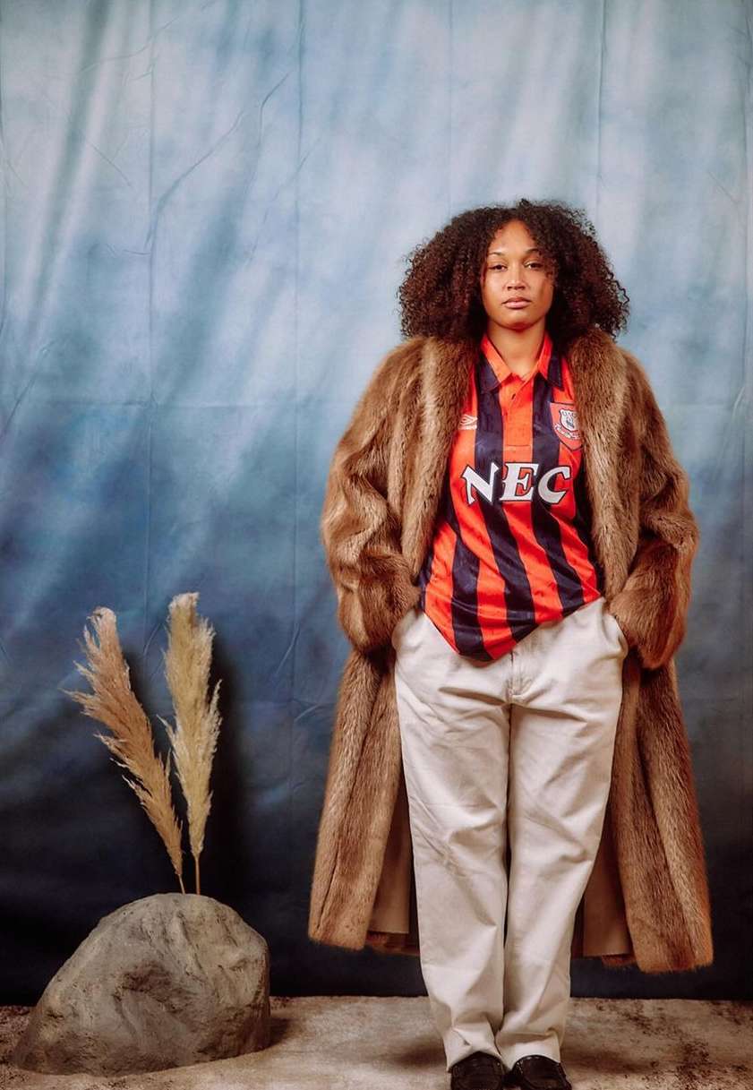 Stylish woman in vintage sports jersey and faux fur coat poses in front of a studio backdrop with stone and wheat accents.