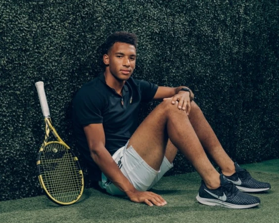 Young man in sportswear sits with a tennis racket against a green wall, wearing a black polo shirt, white shorts, and athletic shoes.