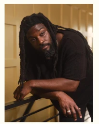 Portrait of a Black man with long dreadlocks and a beard leaning on a railing against a wood panel wall
