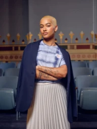 Portrait of a woman with short blonde hair wearing a tie-dye t-shirt, pleated white skirt, and a navy blazer draped over her shoulders.