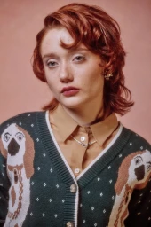 Portrait of a young woman with reddish-brown hair wearing a dark green cardigan with dog pattern and a tan collared shirt by Jared Soares