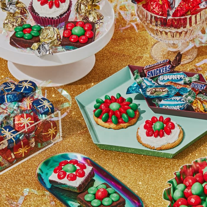 Festive candy treats, including M&M decorated cookies and brownies, wrapped chocolate gifts, and candy in decorative bowls on a gold surface.