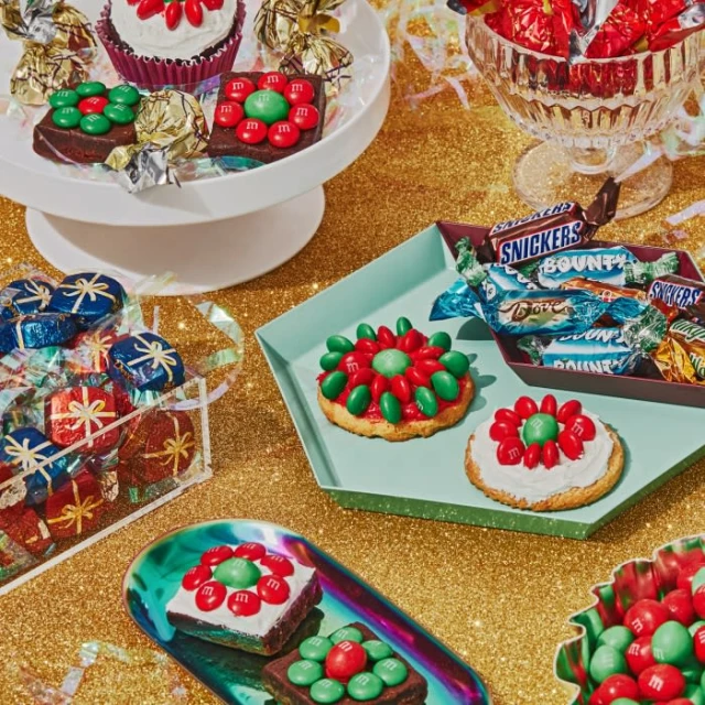 Festive candy treats, including M&M decorated cookies and brownies, wrapped chocolate gifts, and candy in decorative bowls on a gold surface.