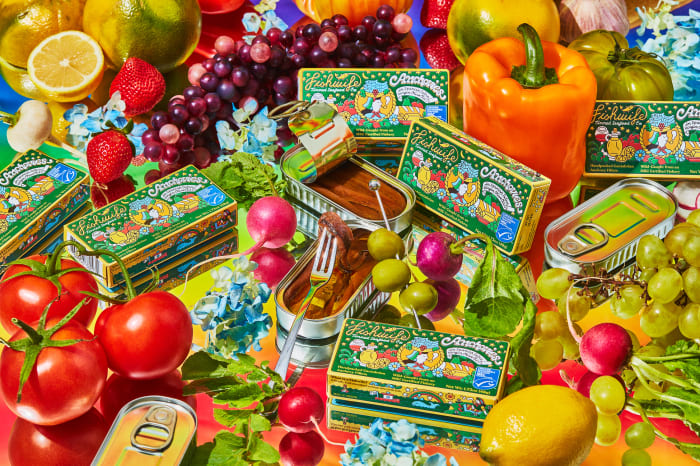 Still life of anchovies in tins surrounded by fresh fruits and vegetables, including tomatoes, grapes, peppers, and lemons, on colorful surface.